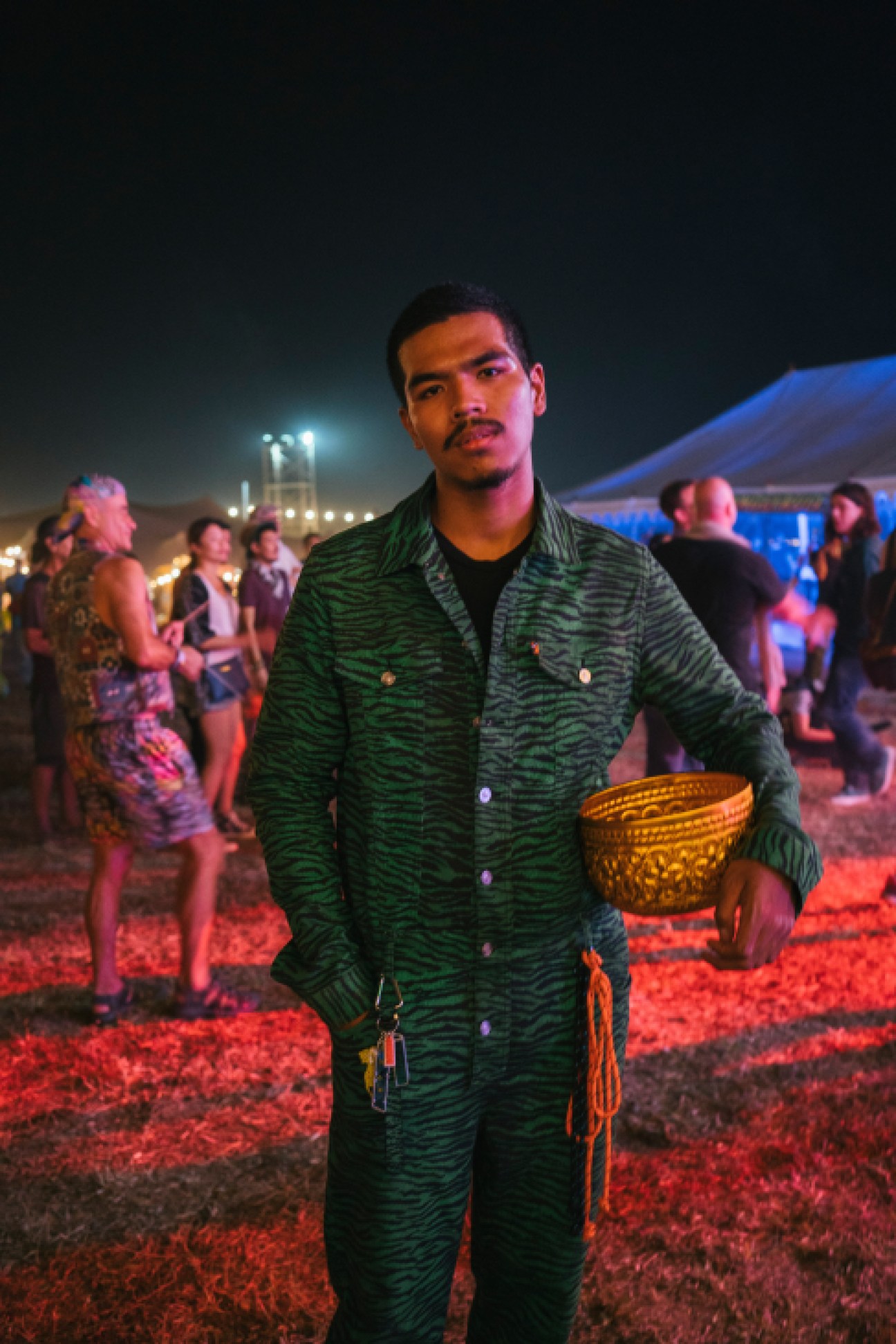Man in tiger-pattern boiler suit holding a golden bowl at Wonderfruit festival at night.