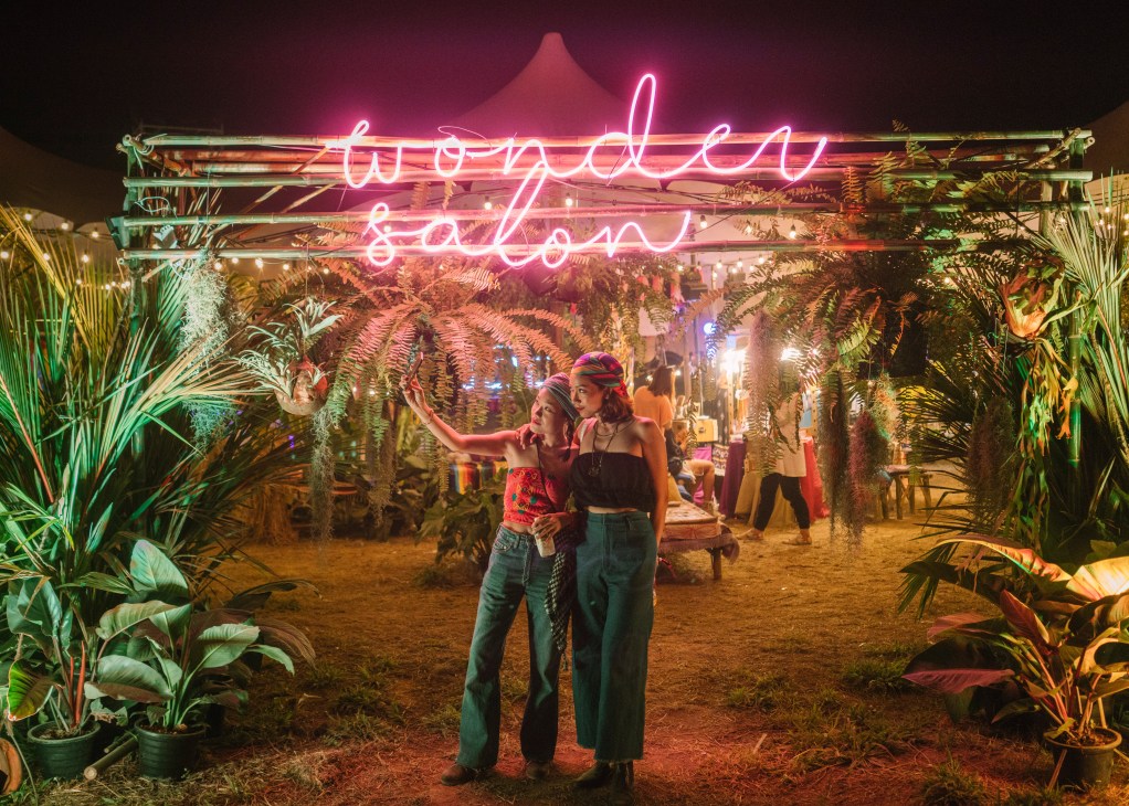 Two people stand under a neon pink sign reading 'wonder salon' in a tropical garden setting at night.