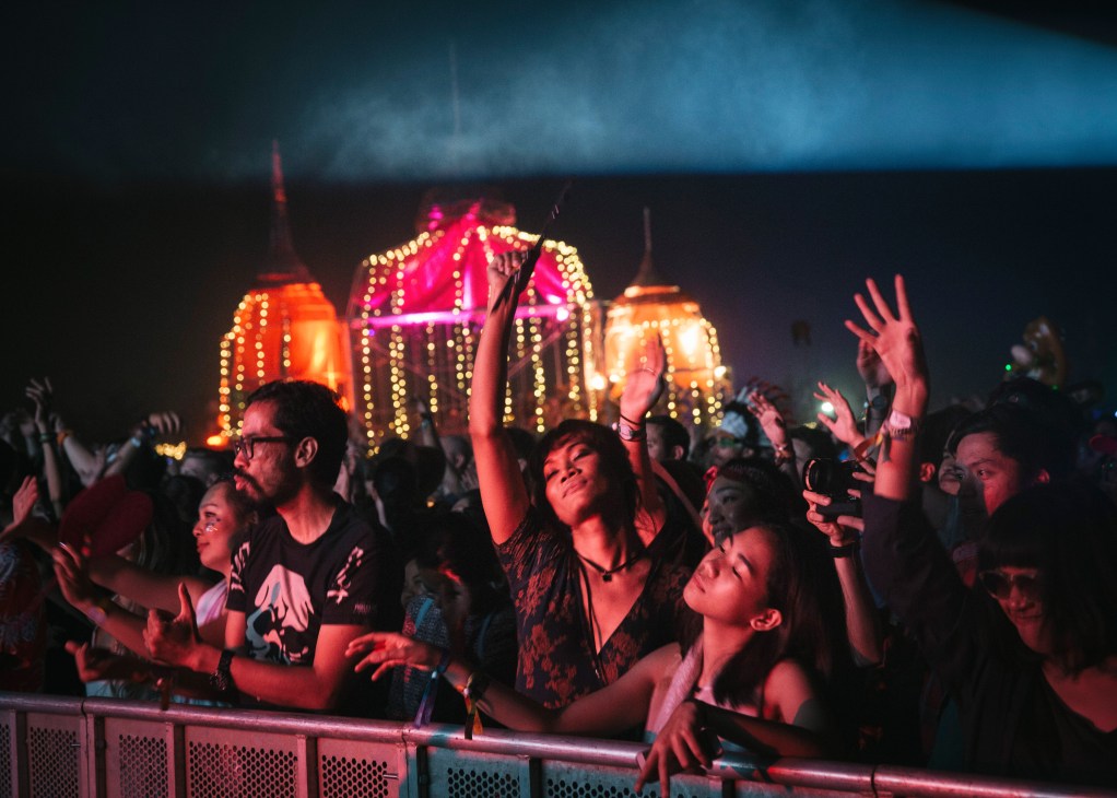 Festival crowd dancing in front of illuminated stage structure at night with blue sky above.