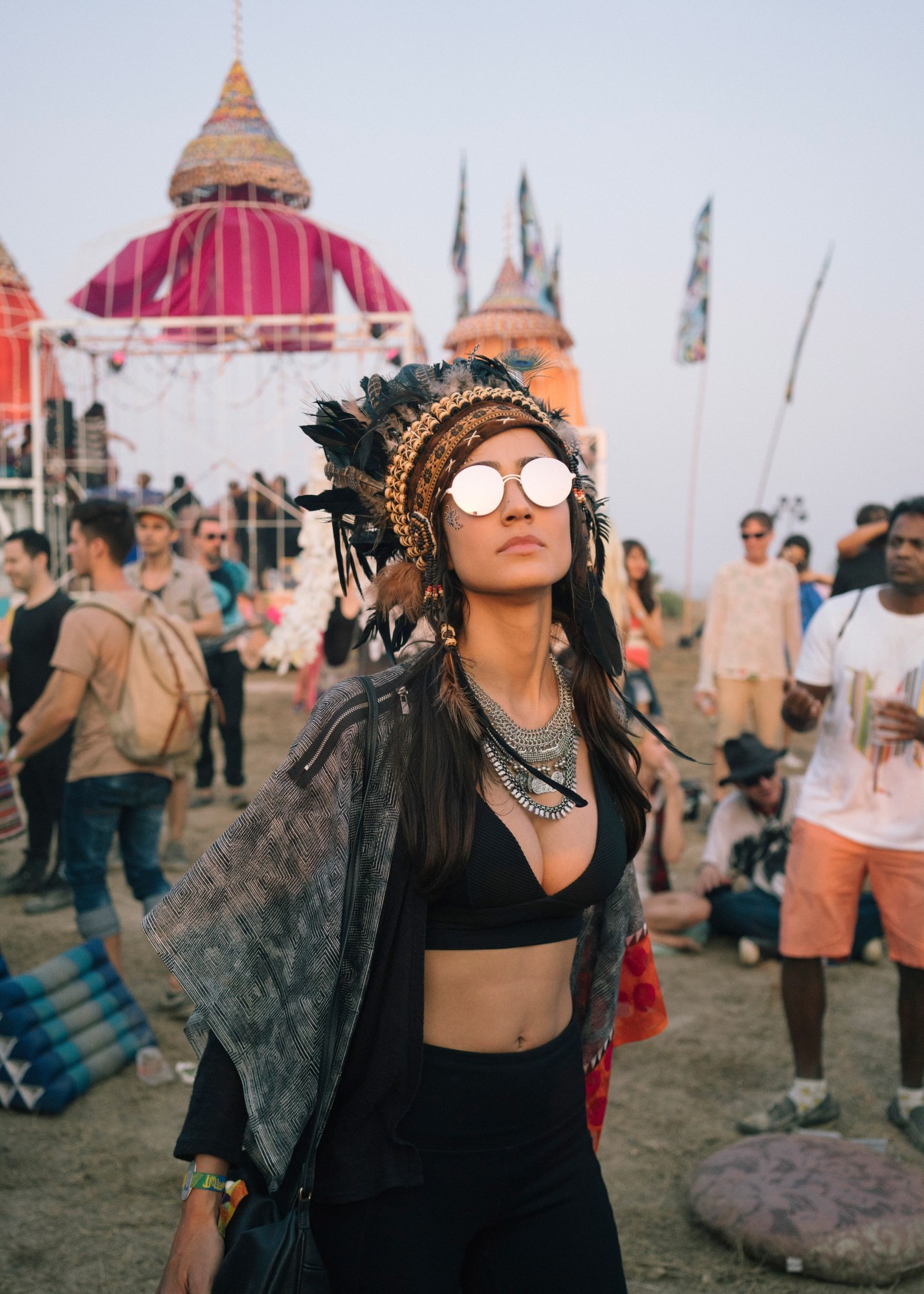 Festival attendee wearing white sunglasses, feathered headdress and ornate jewelry stands before colorful temple-like structures at Wonderfruit festival.