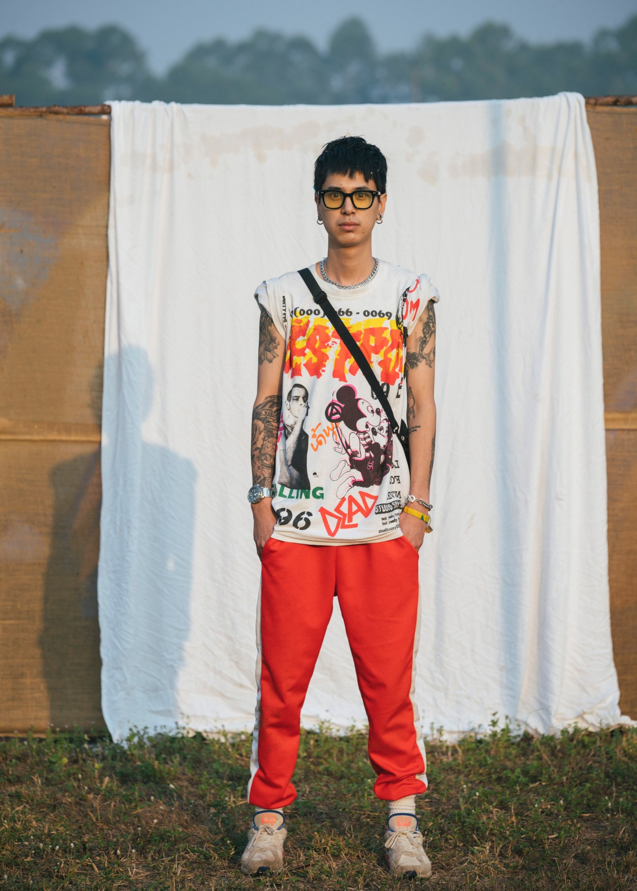 Person wearing graphic print shirt and red pants stands against white fabric backdrop in rural setting.