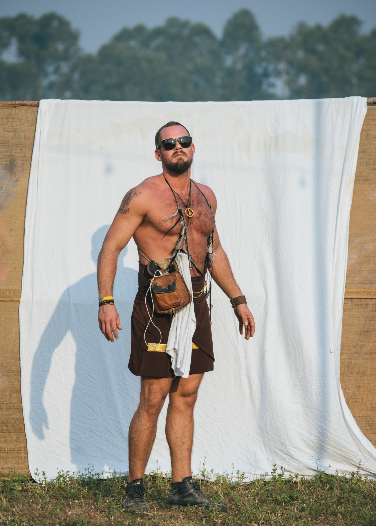 Shirtless man in sunglasses and brown kilt-style skirt posing against white fabric backdrop at Wonderfruit festival.