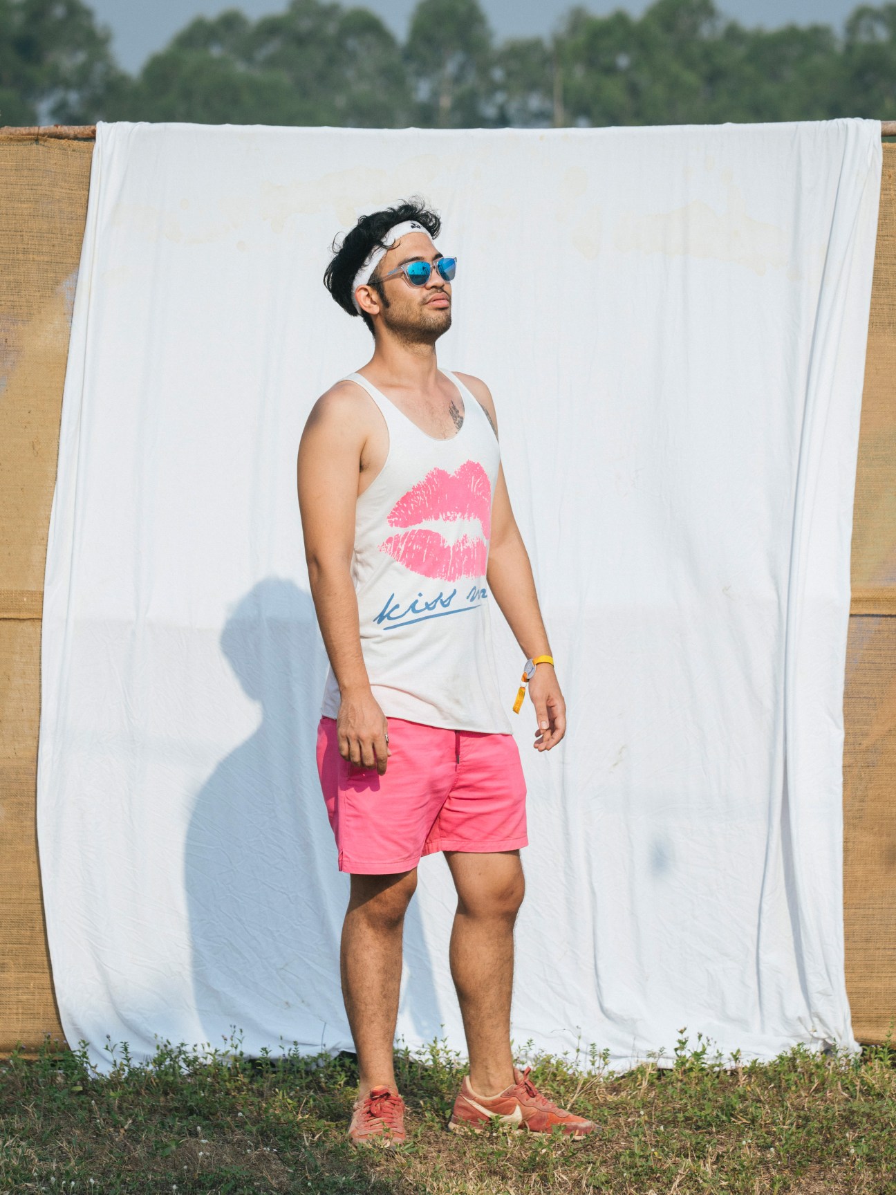 Man in pink shorts and lips-print tank top posing against white fabric backdrop at Wonderfruit festival.