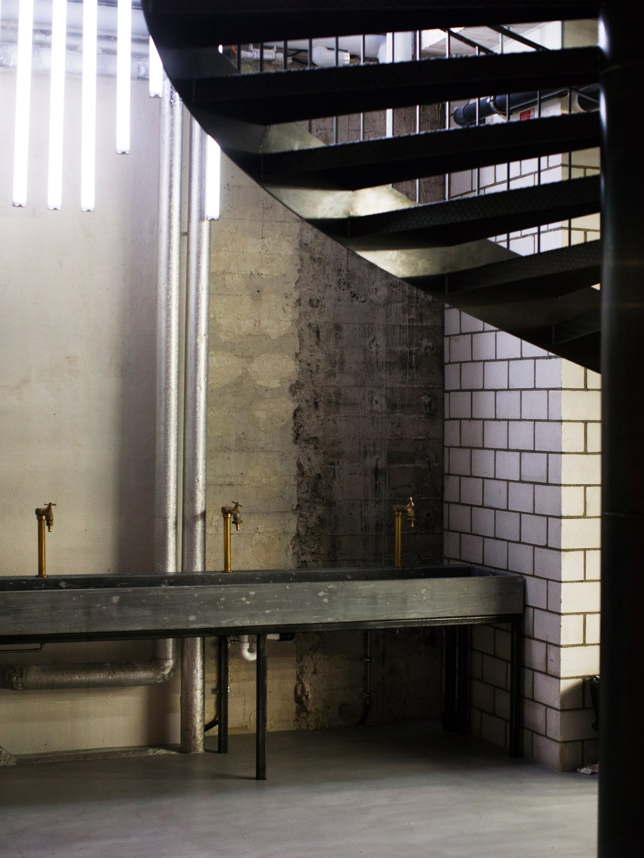 Spiralling black steel stairs descend through an industrial space with exposed brick and concrete walls.