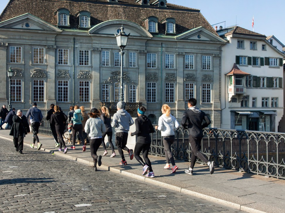 Running club jogging across Münsterbrücke bridge in Zürich past historic architecture.