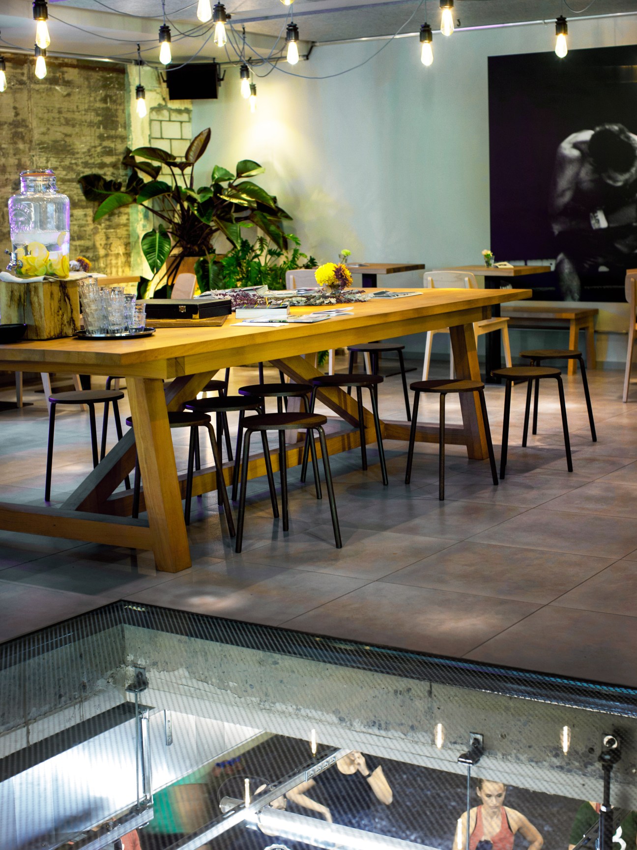 Modern lounge area with wooden table, black chairs, potted plants, and pendant lighting overlooking a gym floor below.