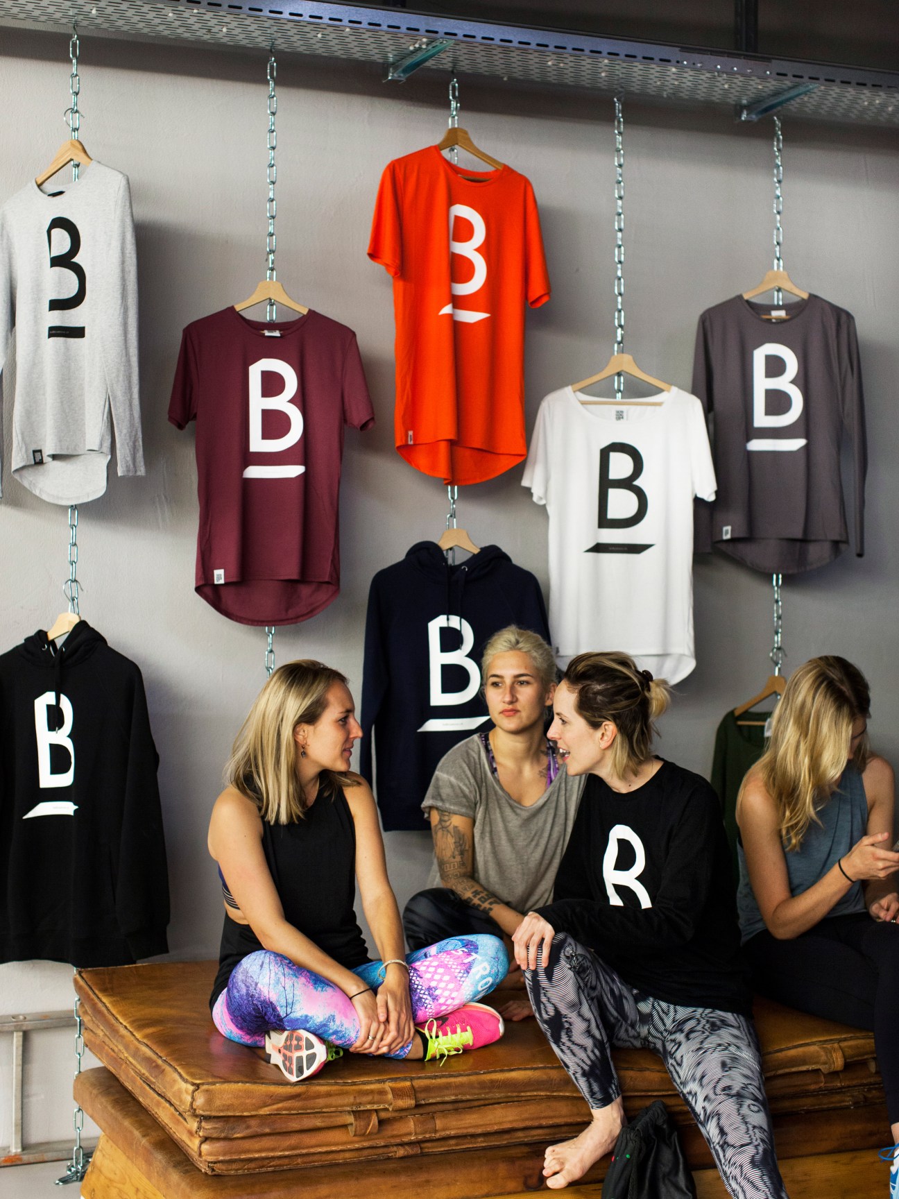 Four people sitting on a wooden bench beneath hanging B-branded t-shirts and hoodies at Balboa Bar & Gym.