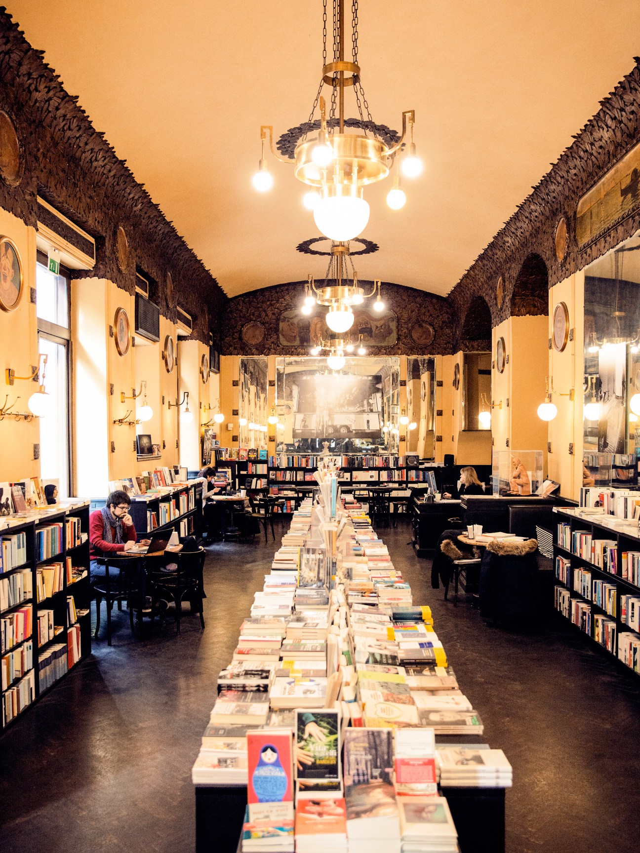 Interior of Caffè San Marco bookshop with yellow walls, hanging pendant lights, bookshelves, and a long table displaying books.