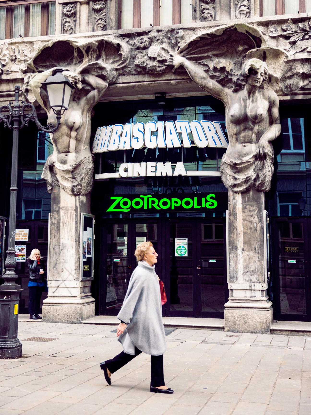 Woman walking past Cinema Ambasciatori entrance with ornate stone caryatid sculptures and neon signs.
