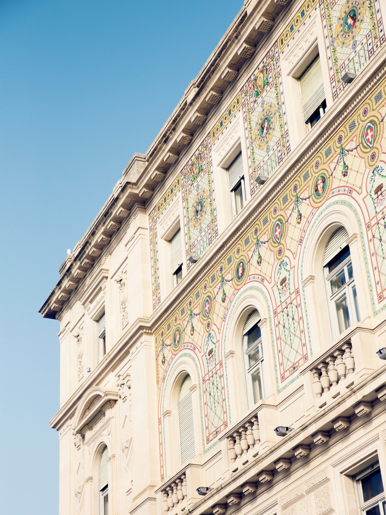 Ornate cream-coloured Austro-Hungarian building facade with colorful mosaic tilework and classical architectural details against blue sky.