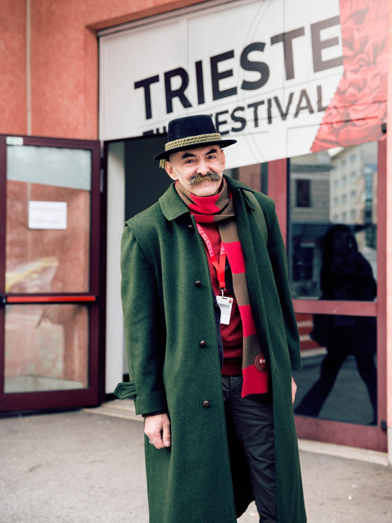 Film critic Ivans Spelkovs stands outside the Trieste Film Festival entrance wearing a green coat, red scarf, and fedora hat.
