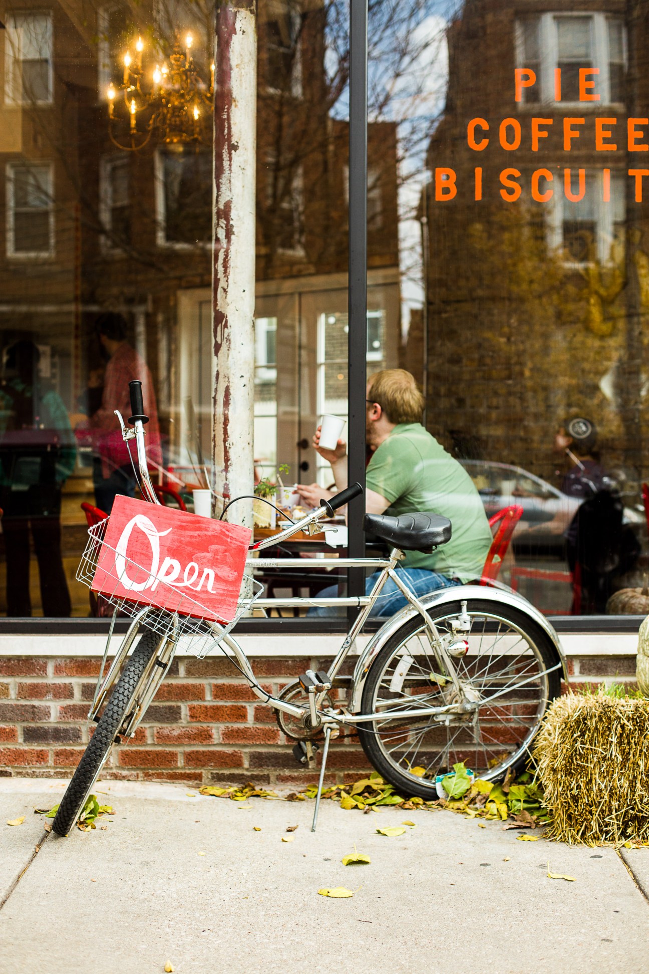 A person in a green shirt sits outside Bang Bang Pie Shop on a bicycle with an open sign in the basket.