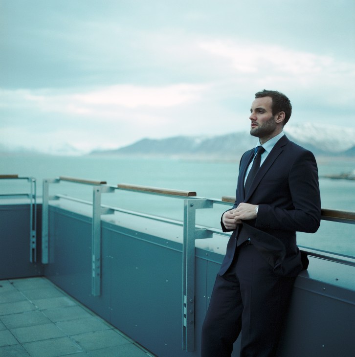 Man in dark suit and tie standing on waterfront balcony overlooking Icelandic bay and mountains.