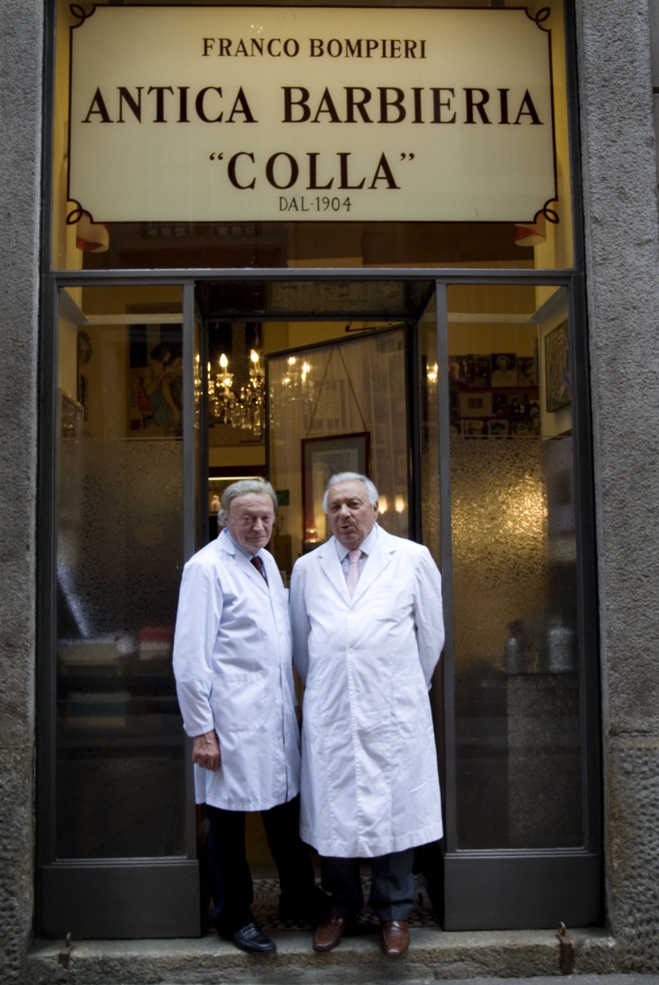 Two men in white barber coats stand in the doorway of Antica Barbieria Colla in Milan.