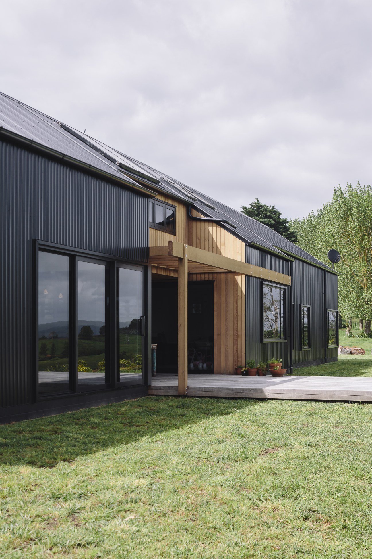 Modern barn-style house with corrugated steel cladding, large windows, and central timber deck extension in New Zealand countryside.