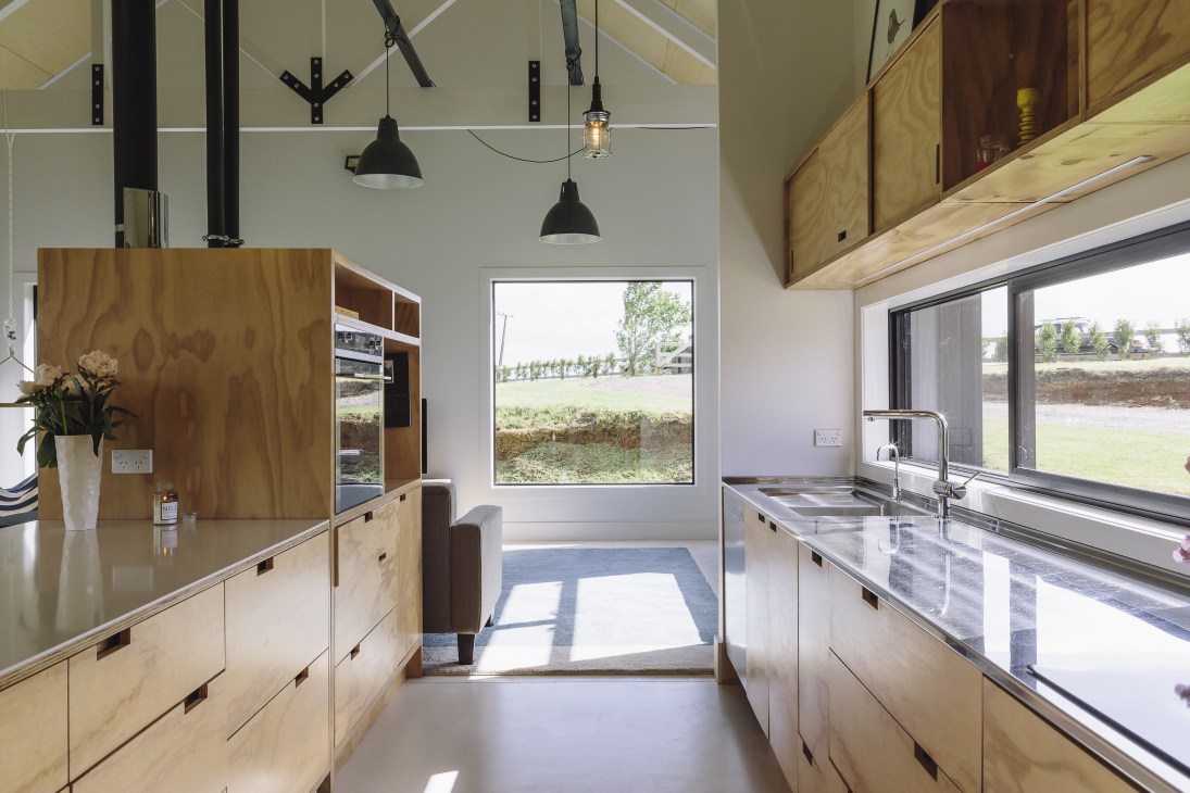 Modern kitchen with plywood cabinetry, pendant lights, and views of rural New Zealand countryside through windows.