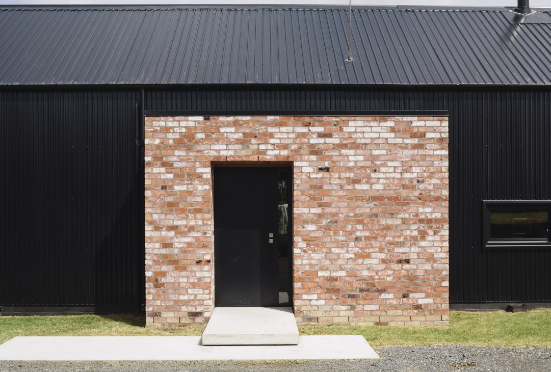 Modern barn entrance with salvaged red brick doorway set against black corrugated steel cladding.