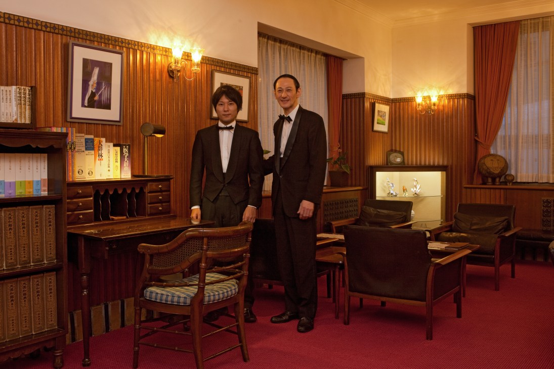 Two bartenders in bow ties stand in a wood-paneled lounge with vintage furniture and framed artwork.