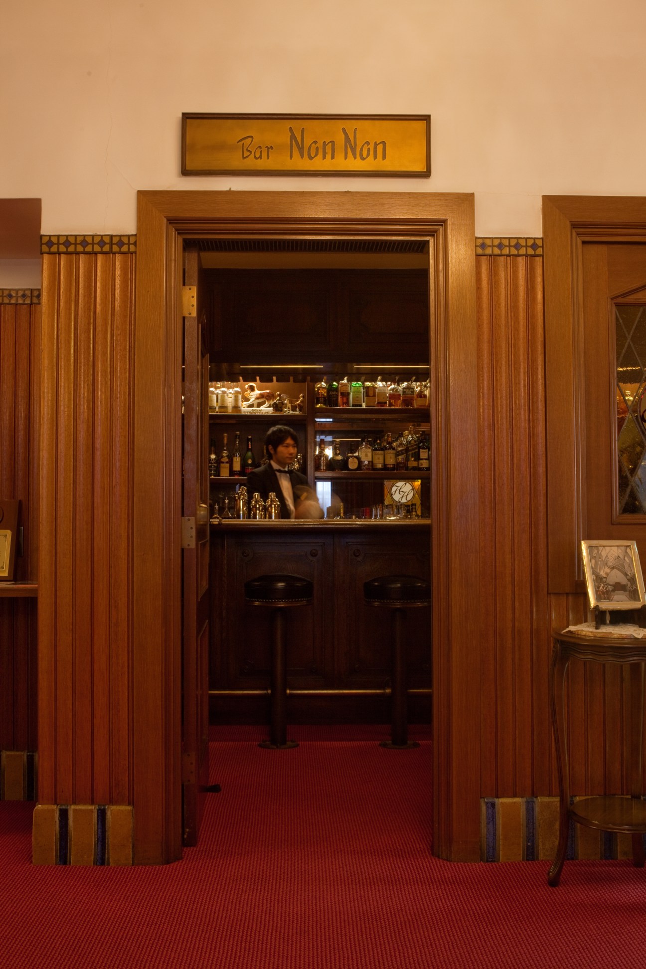 Bar Non Non entrance with wooden signage, framed by polished wood paneling and red carpet, bartender visible behind bar.