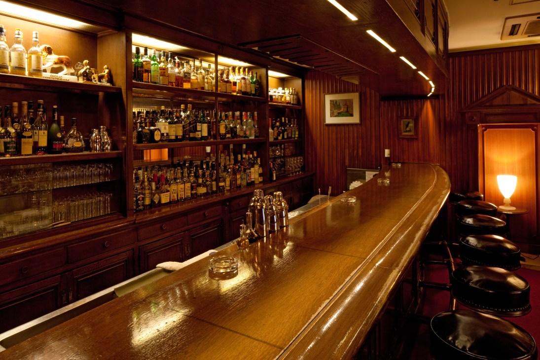 Interior of Bar Non Non showing wooden counter with bar stools, illuminated liquor shelves, and warm lighting.