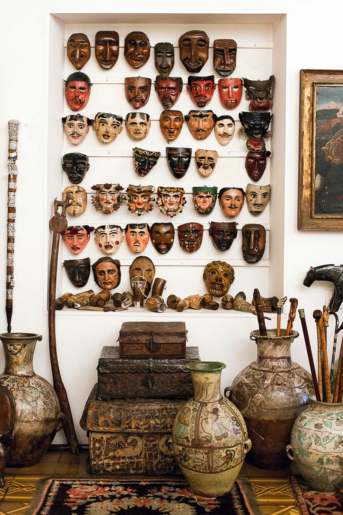 Collection of Peruvian masks and artifacts displayed on white shelves with ceramic vessels and tools below.