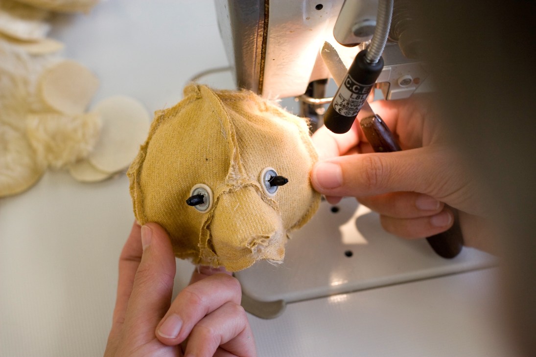 Steiff worker hand-stitching a teddy bear's head with glue applicator at sewing machine.