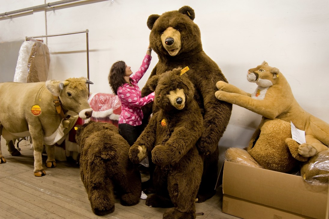 A Steiff employee adjusts a large brown teddy bear surrounded by other oversized plush animals in a workshop.