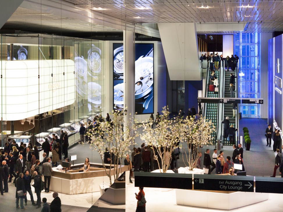 Baselworld exhibition hall with attendees, decorative flowering branches, illuminated displays, and escalators.