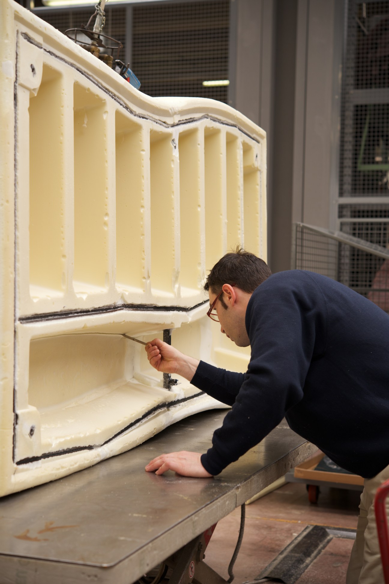Worker trims excess polyurethane foam from a large cream-colored furniture piece using a handheld tool.