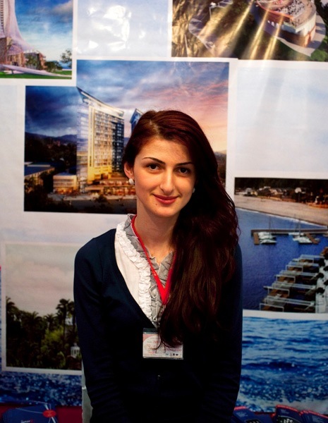 Woman vendor at Batumi Expo standing in front of photographs of Batumi development projects.