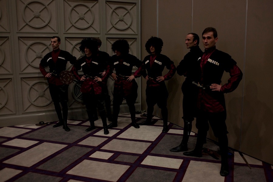 Six performers in traditional Georgian black and red costumes with decorative medallions stand posing against a paneled wall.