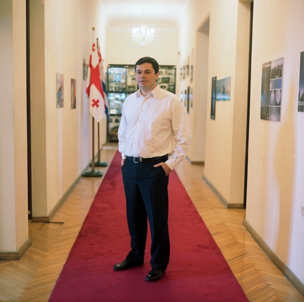 Man in white shirt and dark trousers standing on red carpet in hallway with Georgian flag visible