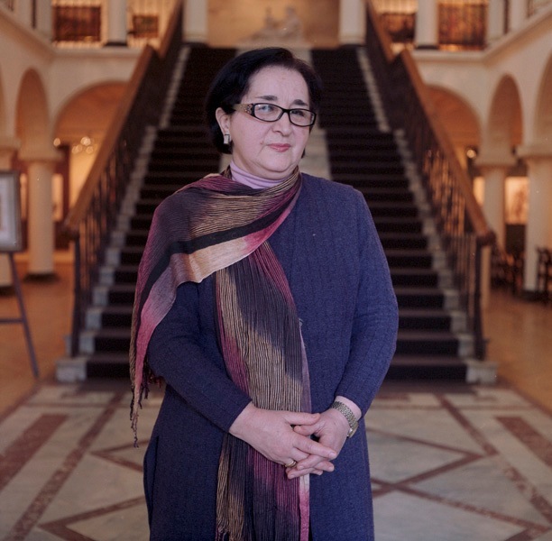 Woman with glasses wearing a dark blue sweater and patterned scarf stands in an ornate interior with a grand staircase behind her.