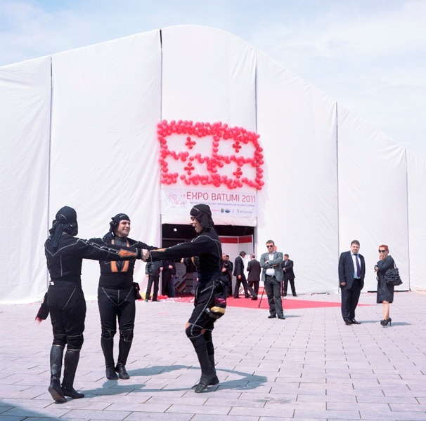 Traditional dancers in black costumes perform outside a large white tent structure with pink lettering for Expo Batumi 2011.