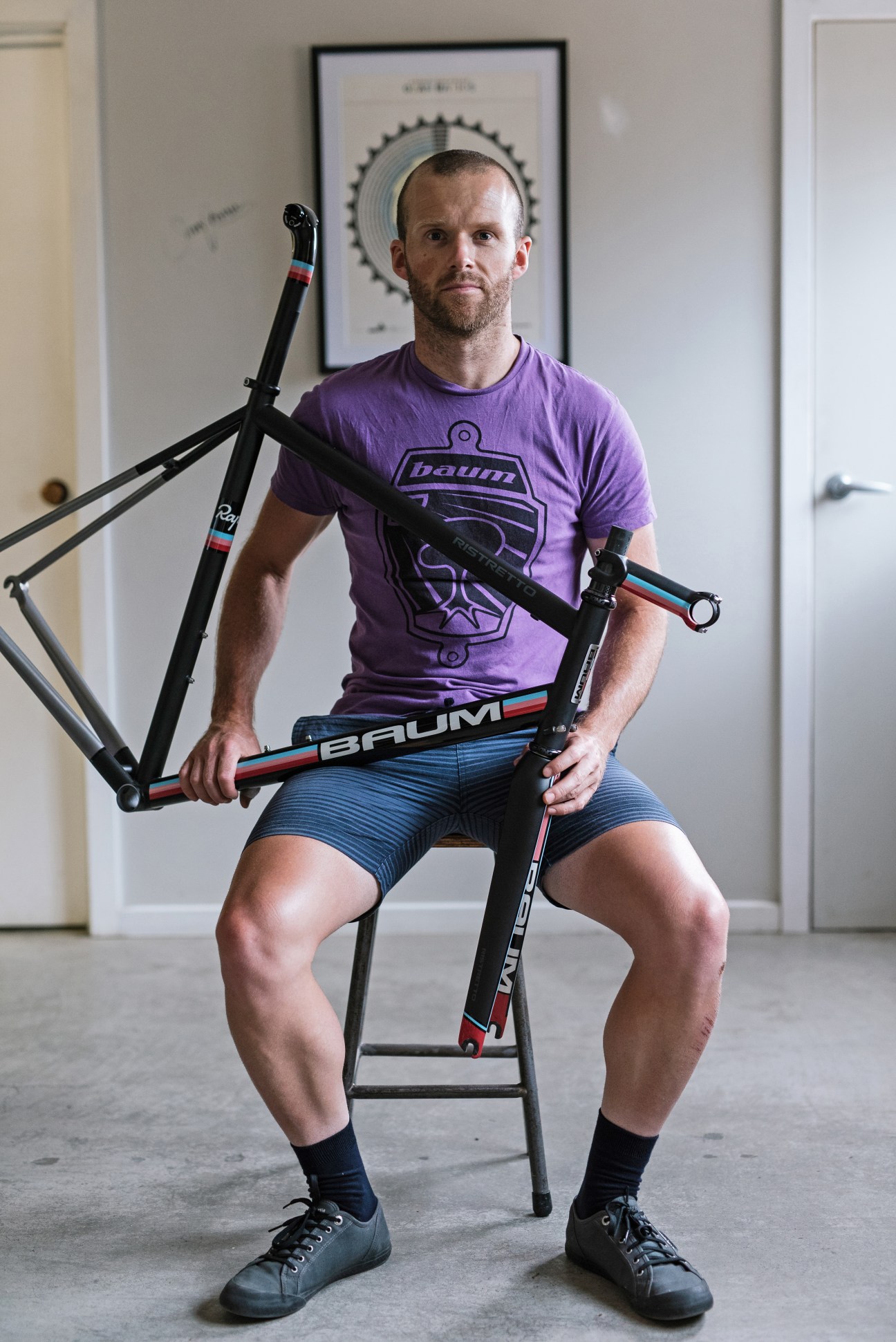 Man in purple Baum t-shirt holding handcrafted bicycle frames in workshop.