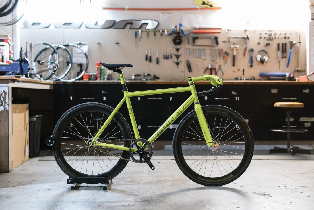 A finished lime-green Baum Cycles track bike in a workshop with tools and bike wheels on pegboard wall.