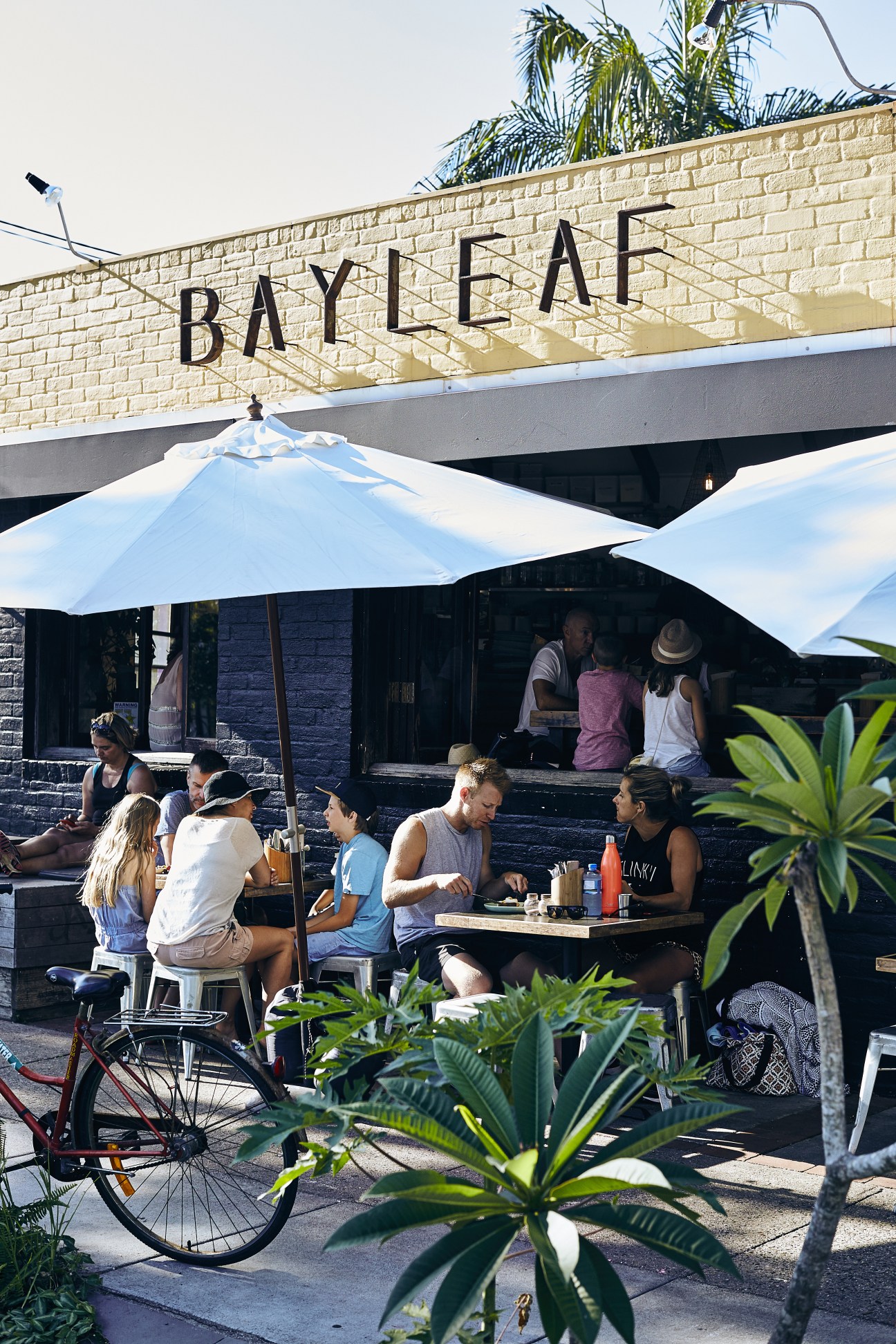 Patrons dining at outdoor tables under umbrellas at Bayleaf café in Byron Bay, with signage visible on the brick building.