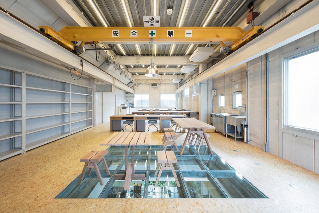 Spacious workshop interior with yellow overhead crane, wooden workbenches, storage shelving, and glass floor section revealing lower level.