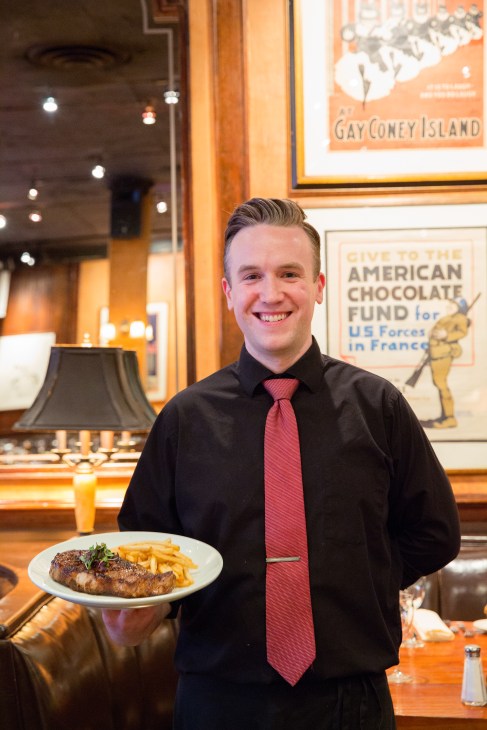 A smiling waiter in a black shirt and red tie holds a plate of steak and chips at the Knickerbocker restaurant.