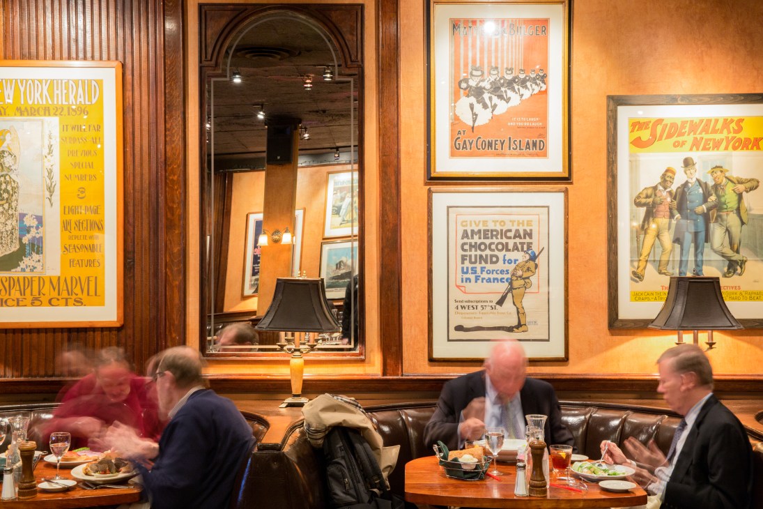 Interior of Knickerbocker restaurant showing wood-panelled walls, vintage posters, and diners at leather banquettes.