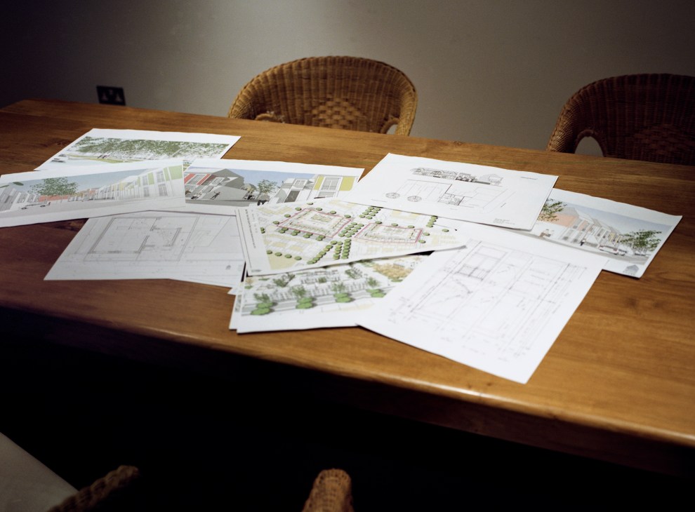 Architectural plans and sketches for the Rational House displayed on a wooden table with wicker chairs.