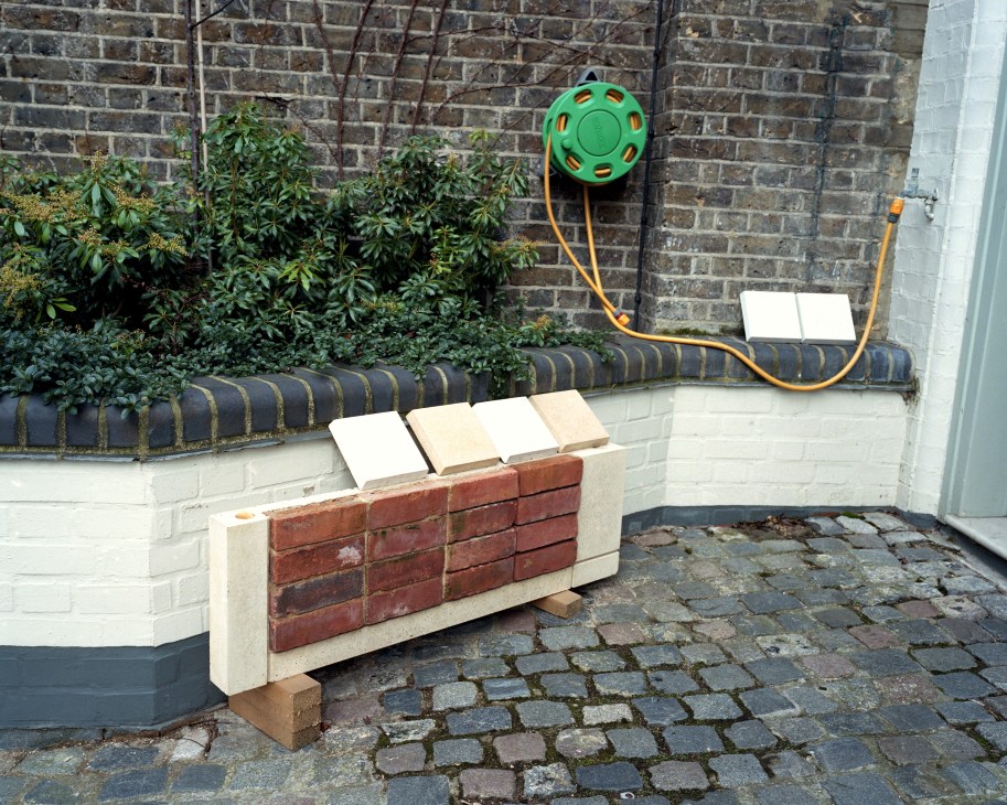 Material samples of brick and concrete displayed on a bench against a brick wall with a garden hose.