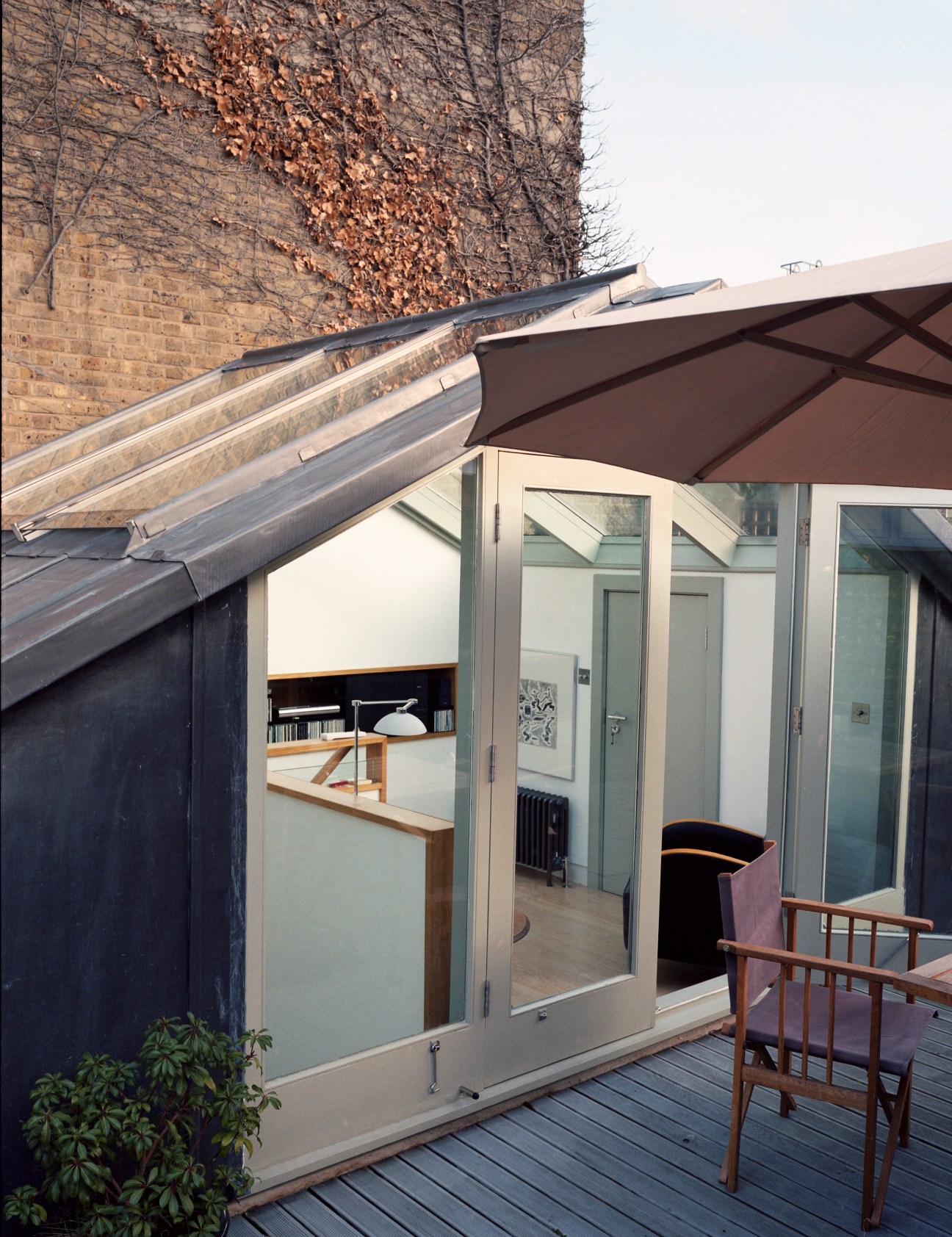 Roof terrace with wooden decking, modern glass doors, brown umbrella, wooden chair, and climbing ivy on brick wall.