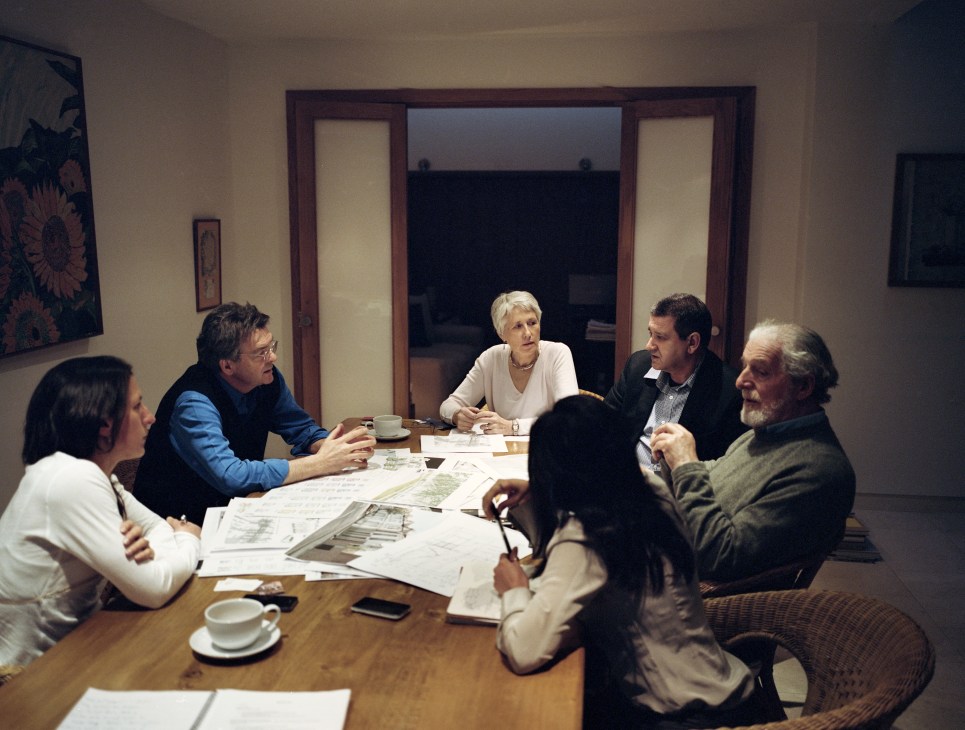 Six people seated around a wooden table reviewing architectural plans and documents during a meeting.