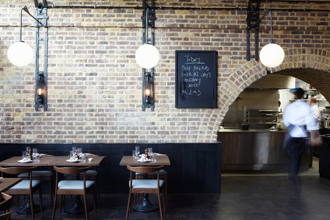 Restaurant interior with exposed brick walls, pendant lights, wooden tables and chairs, and an open kitchen visible through an arched opening.