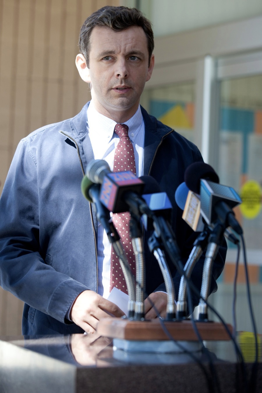 Man in blue jacket and burgundy tie speaking at microphones during press conference.