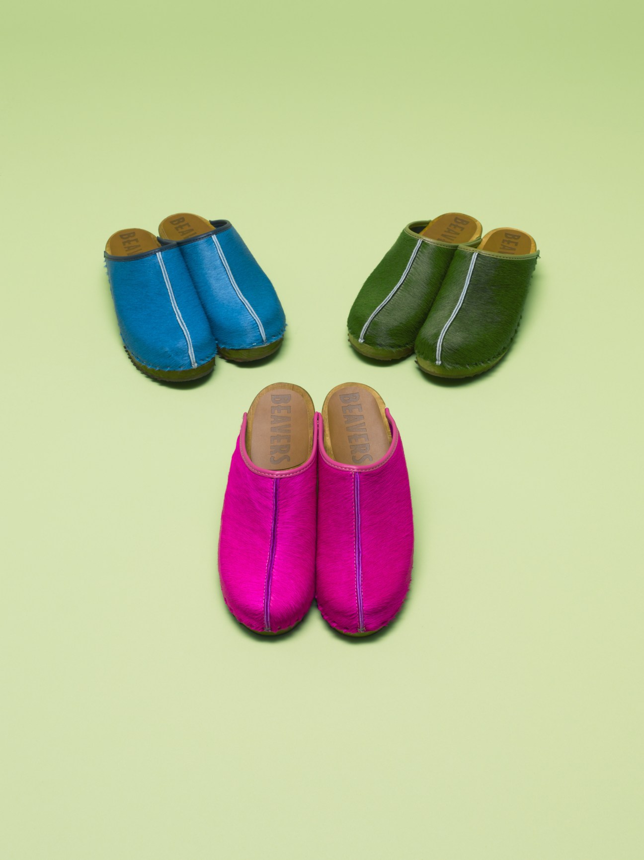 Three pairs of colorful Beavers clogs in blue, green, and magenta with wooden soles on a pale green background.