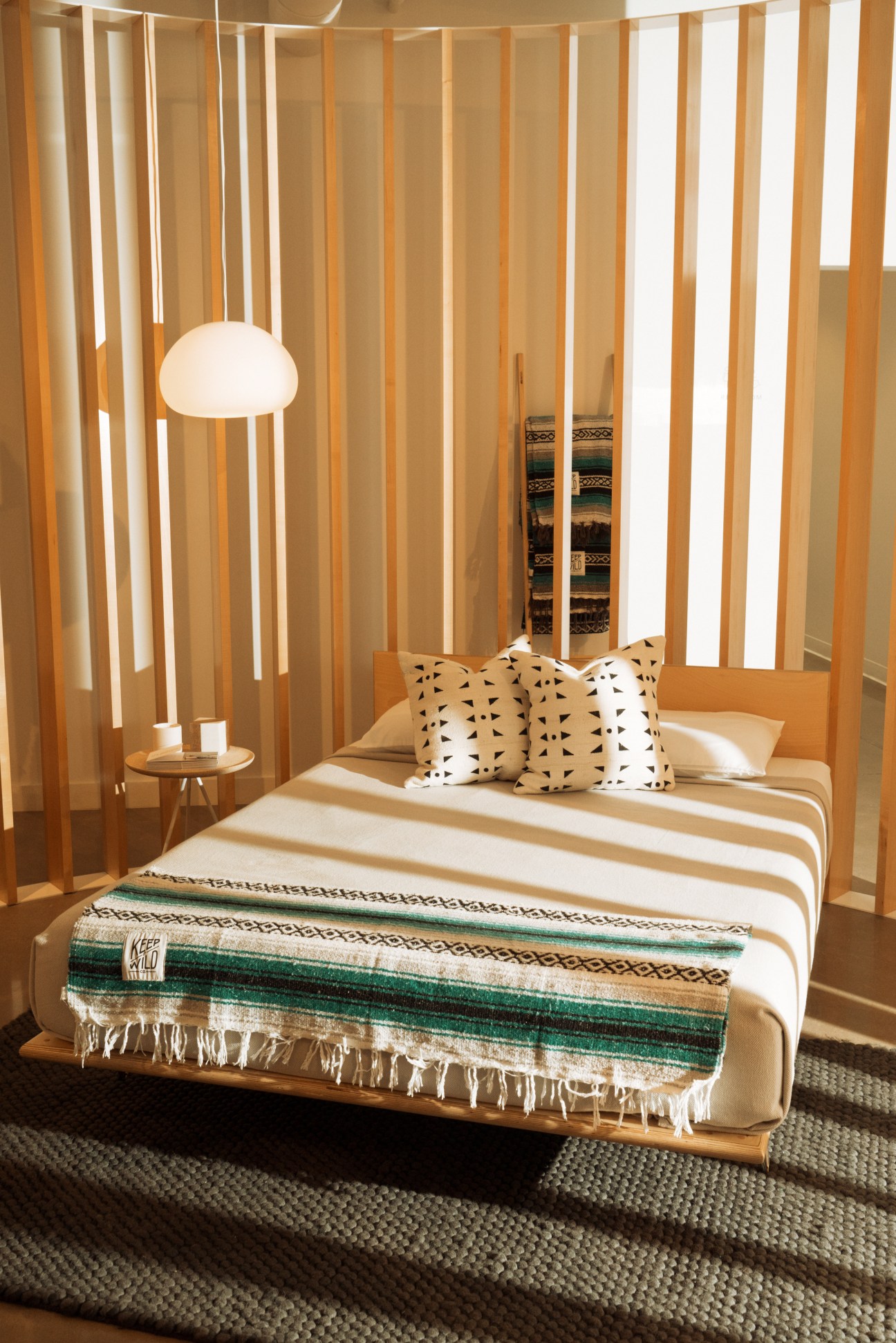 Wooden bed frame with striped mattress, patterned pillows, and hanging pendant lamp in modern bedroom display.