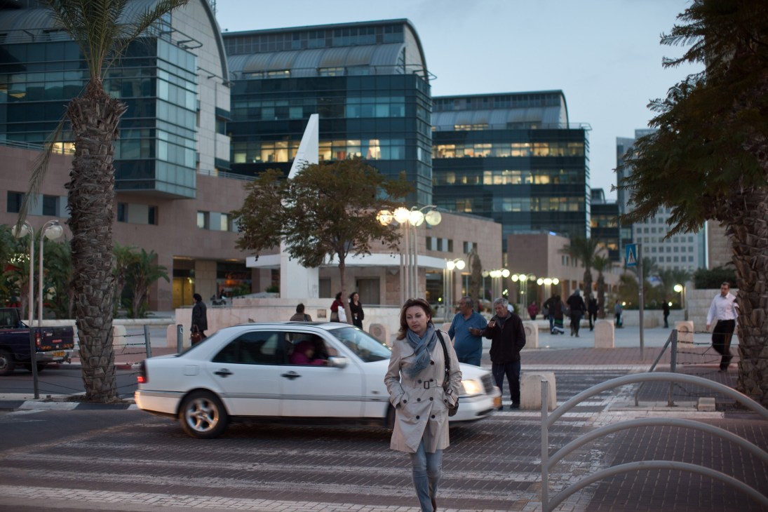 Woman in beige trench coat standing in Be'er-Sheva city center at dusk with modern glass buildings and palm trees.