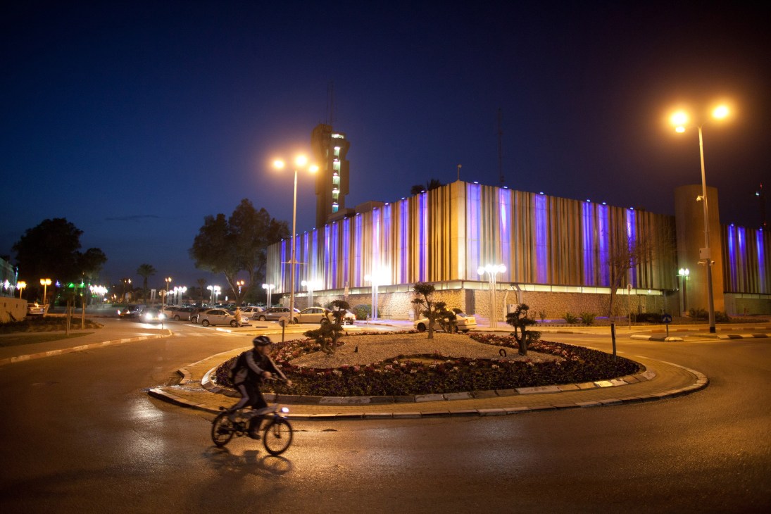 Person cycling past illuminated modern building with purple and gold vertical striping at dusk in Be'er-Sheva.